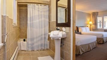 Une salle de bain d’hôtel avec une douche, un lavabo et un miroir. La pièce adjacente contient deux lits.