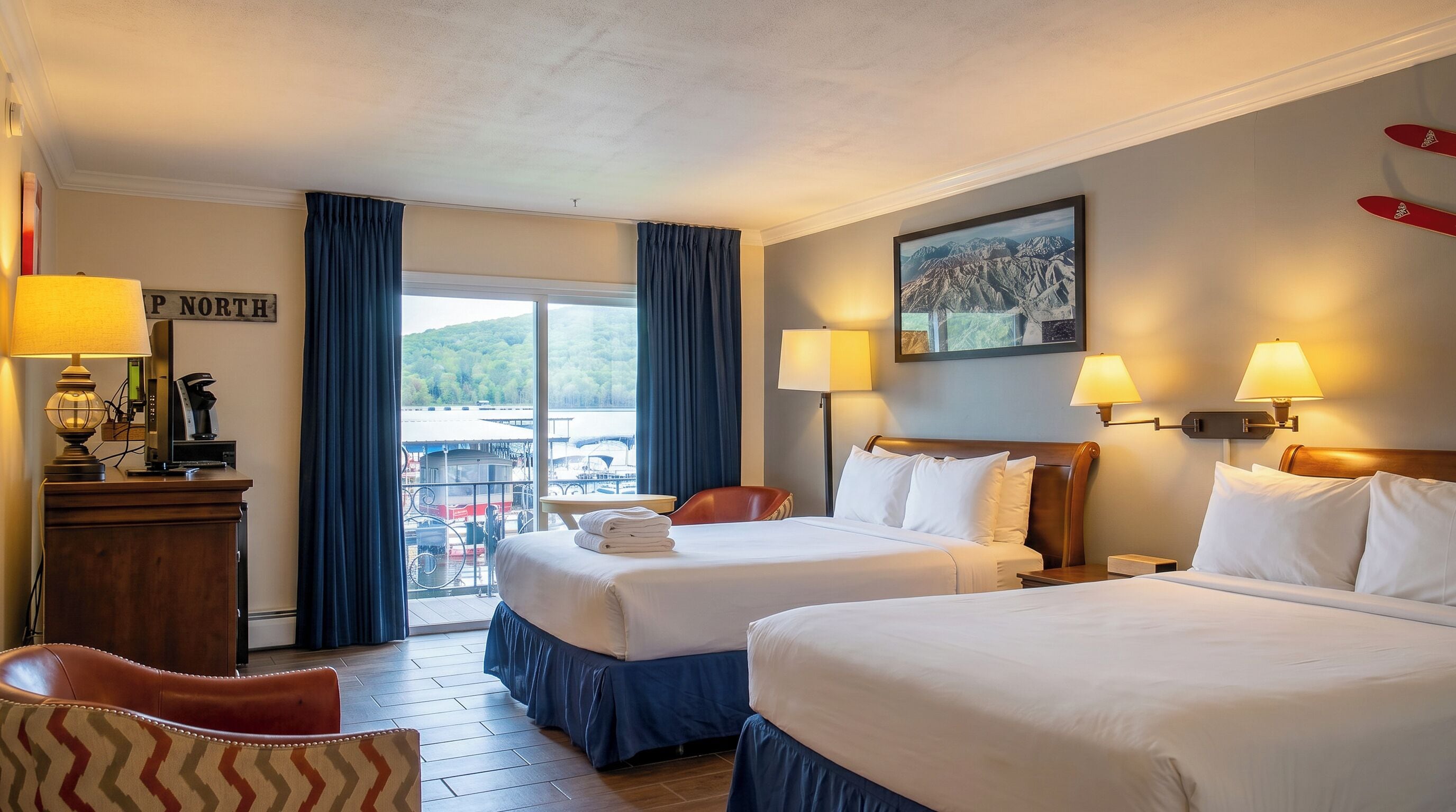 A hotel room with two beds, a desk with a lamp, a chair, and a view of a dock with boats.