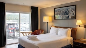 A hotel room with a bed, two bedside lamps, a chair, a small table, and a large window with a view of boats and mountains. - Gauthier's Saranac Lake Inn (Saranac Lake)