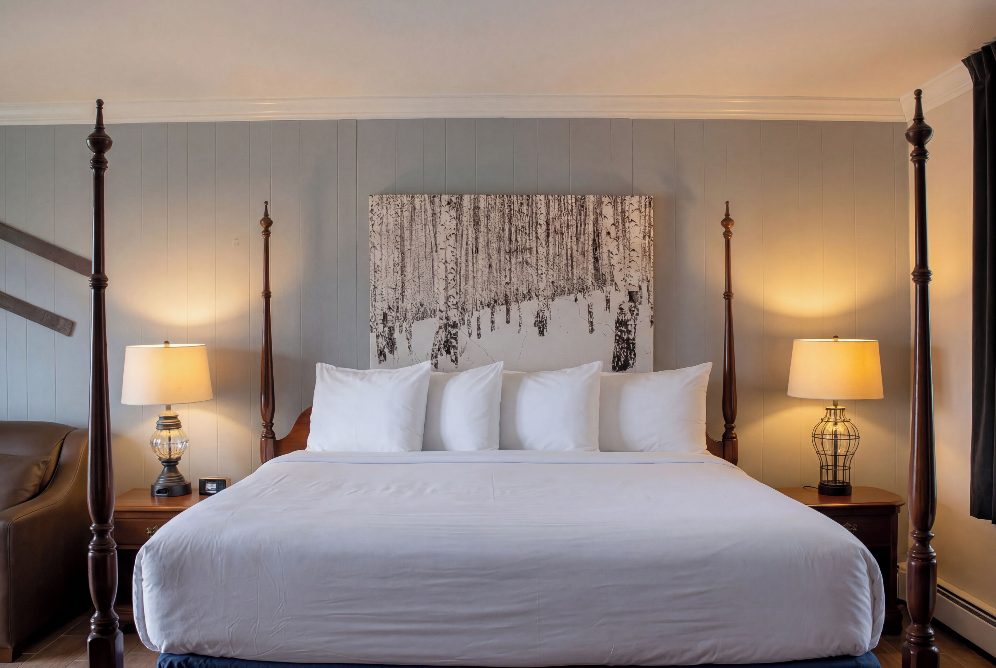 A large bed with white linens, two bedside tables with lamps, a headboard with a hanging artwork, and a wooden chair.
