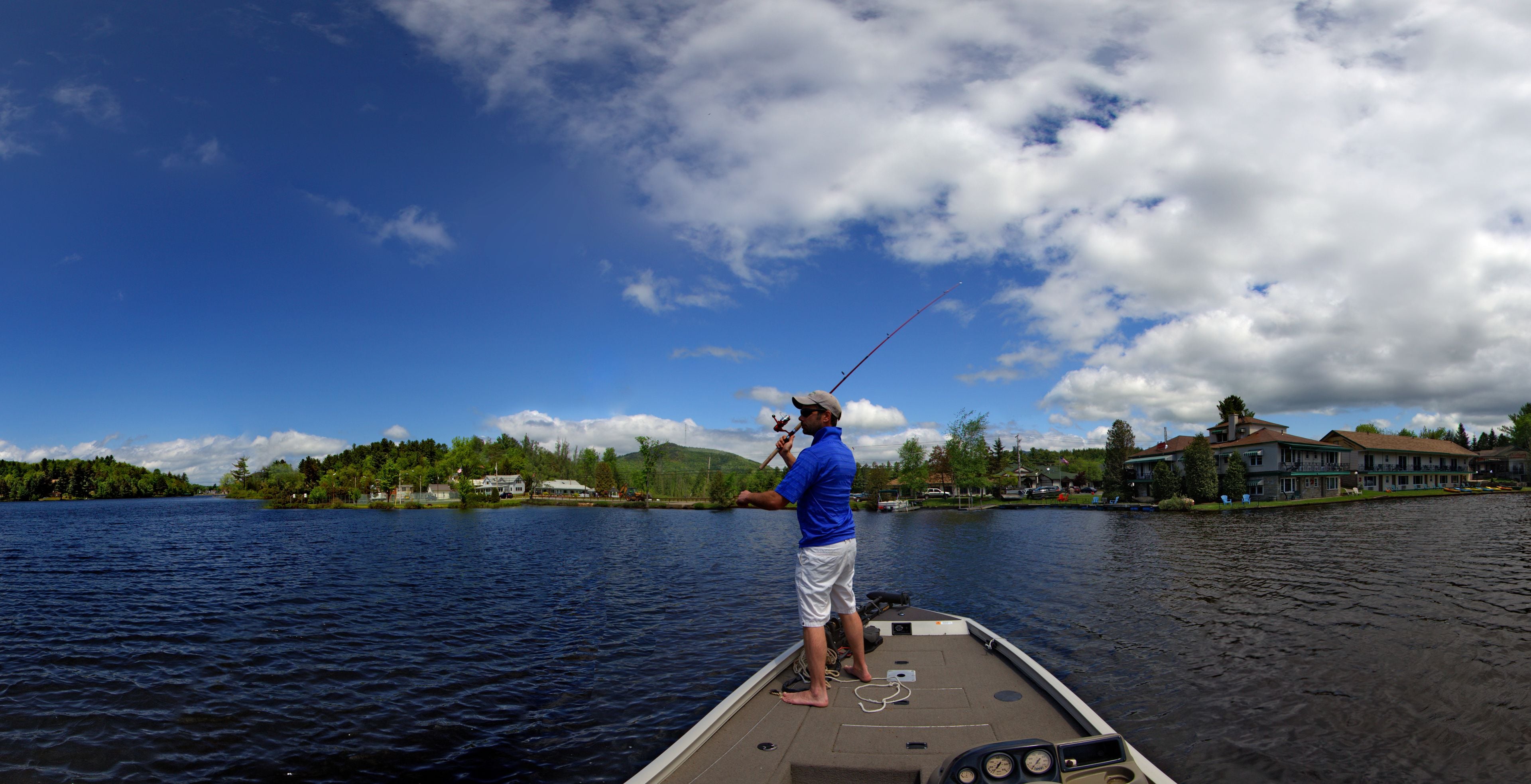 A person fishing on a boat with a house and trees in the background.