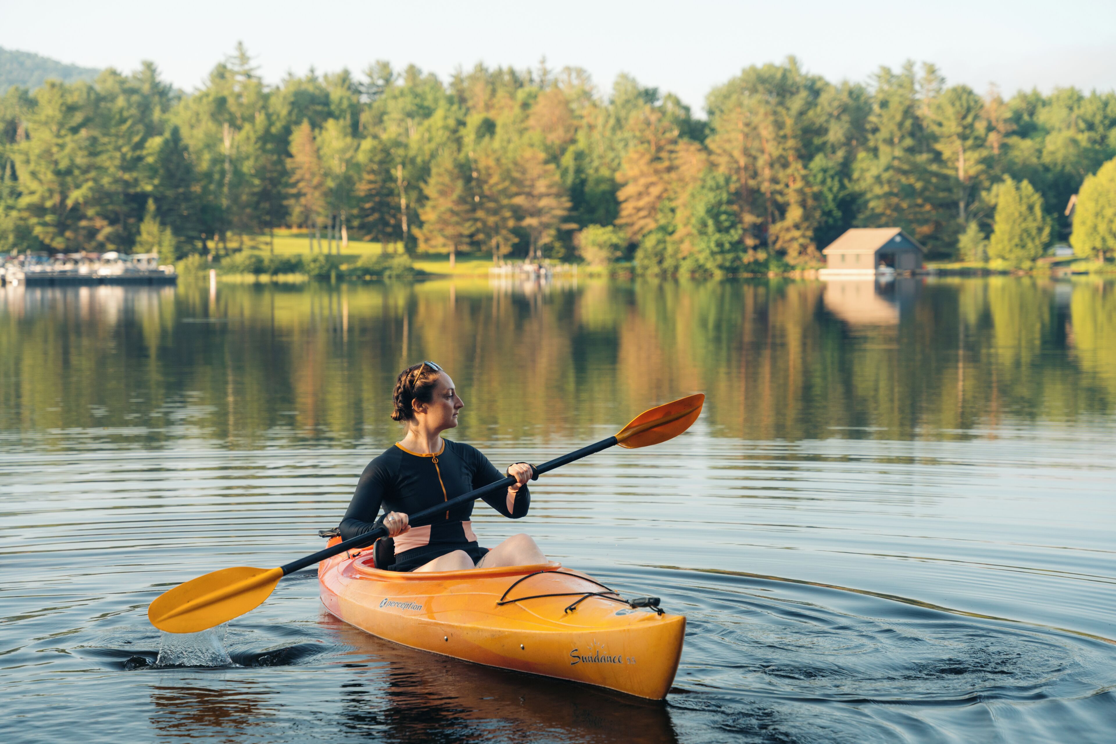 Una persona haciendo kayak en un lago tranquilo, con árboles y una cabaña al fondo.