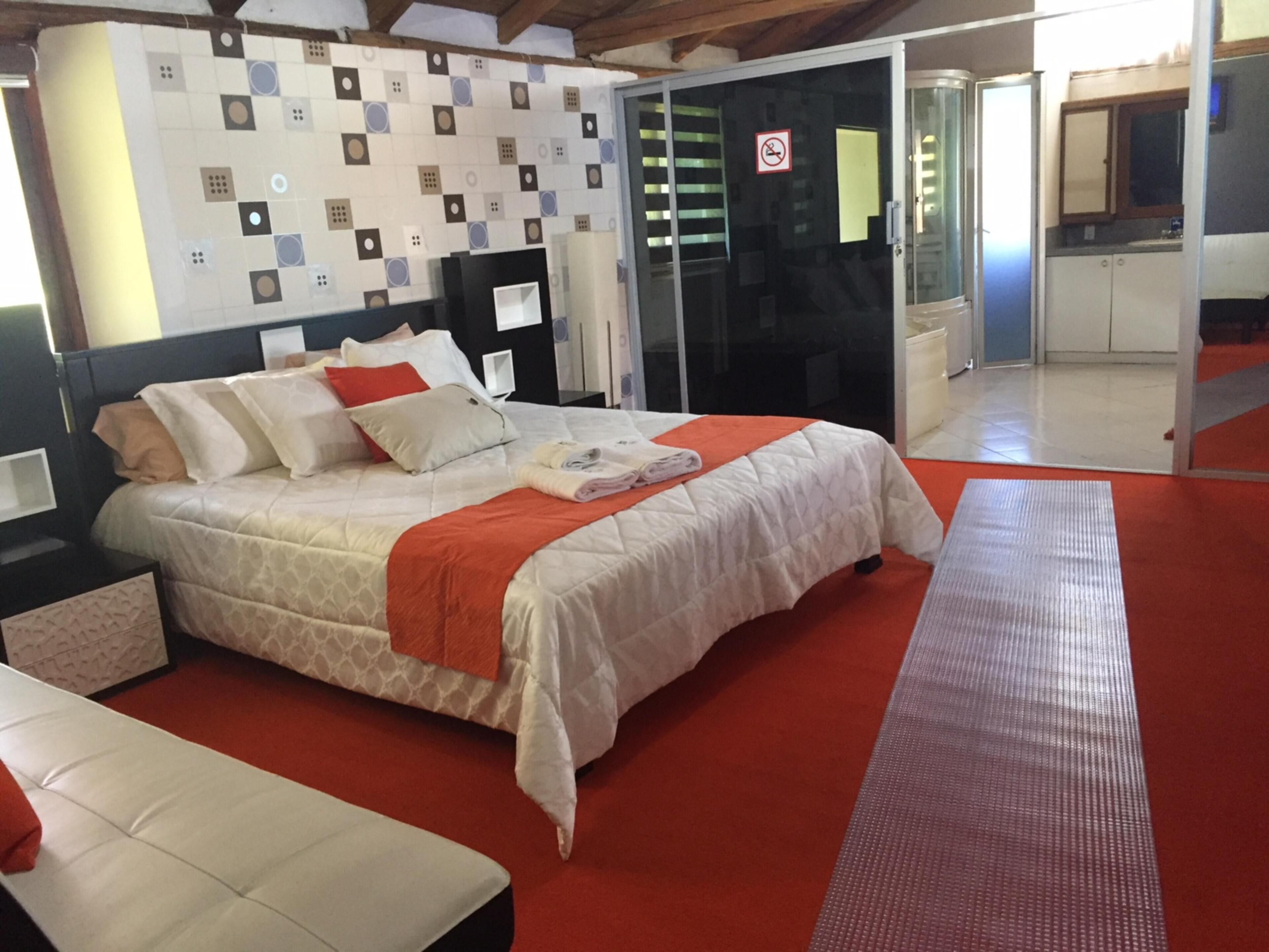 Una habitación de hotel ordenada con una cama, una mesita de noche pequeña, una mesita auxiliar, una alfombra roja y una pared con un diseño de baldosas geométricas.