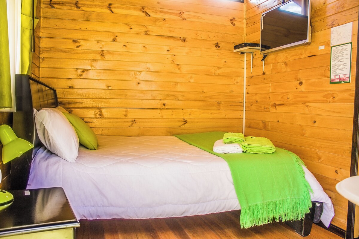 Una cama individual con una manta verde, un televisor montado en la pared y un panel de pared de madera.