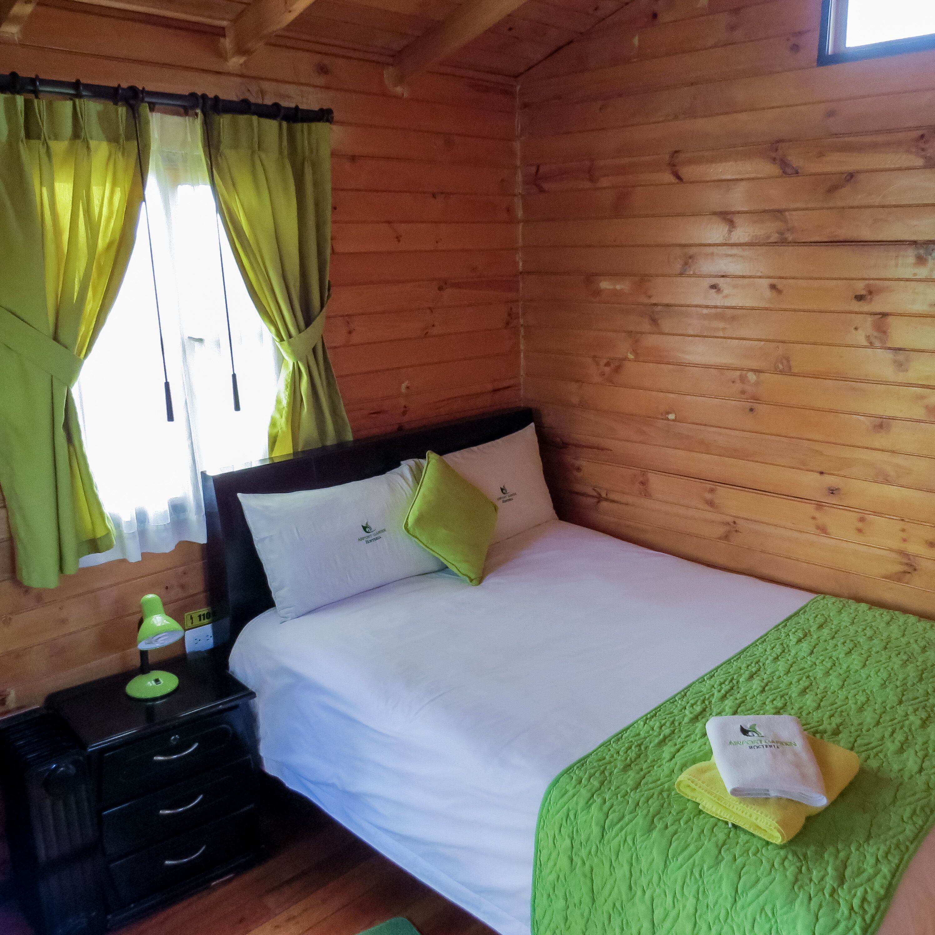 Habitación tipo cabaña de madera con una cama, una mesita de noche, una lámpara y una ventana con cortinas.