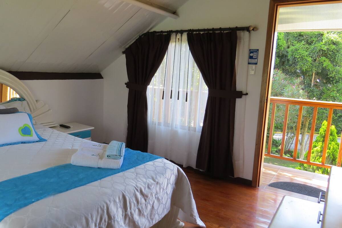 Un dormitorio con cama, mesita de noche, ventana con cortinas y vista a zonas verdes.