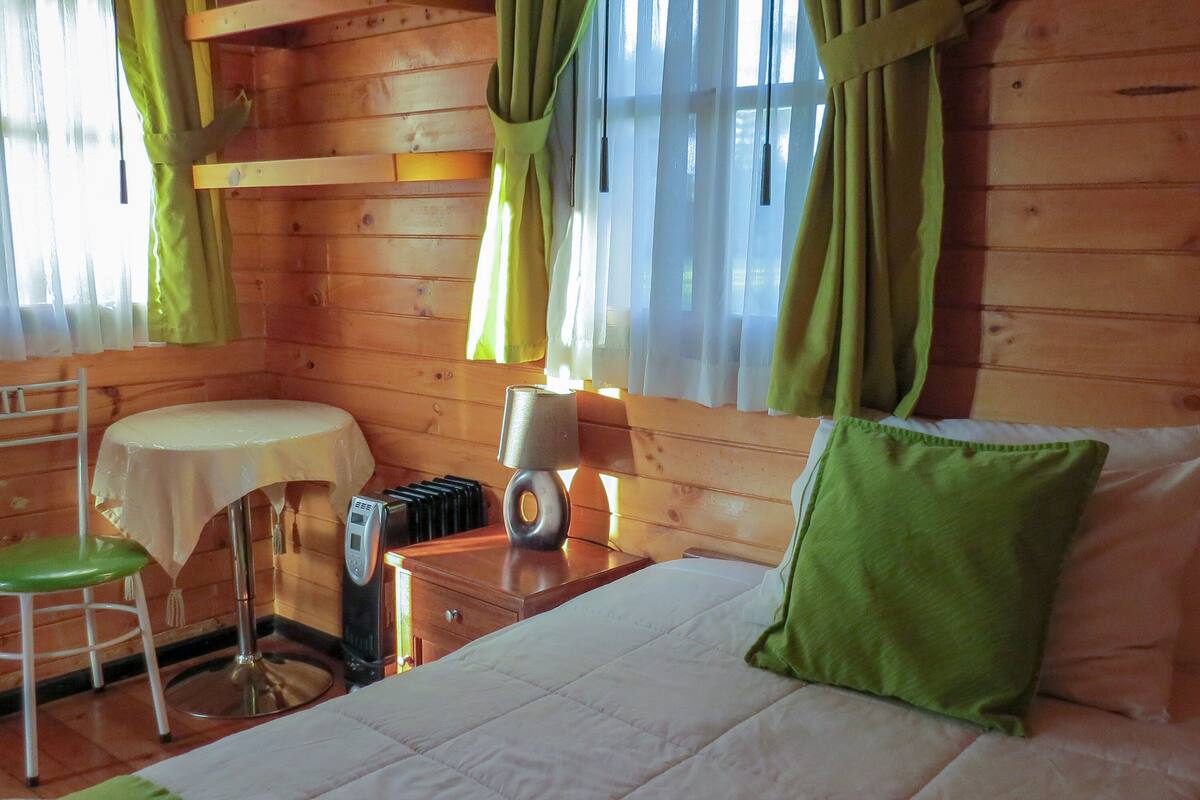 Una habitación de cabaña de madera con una cama, una mesa, una silla y una lámpara.