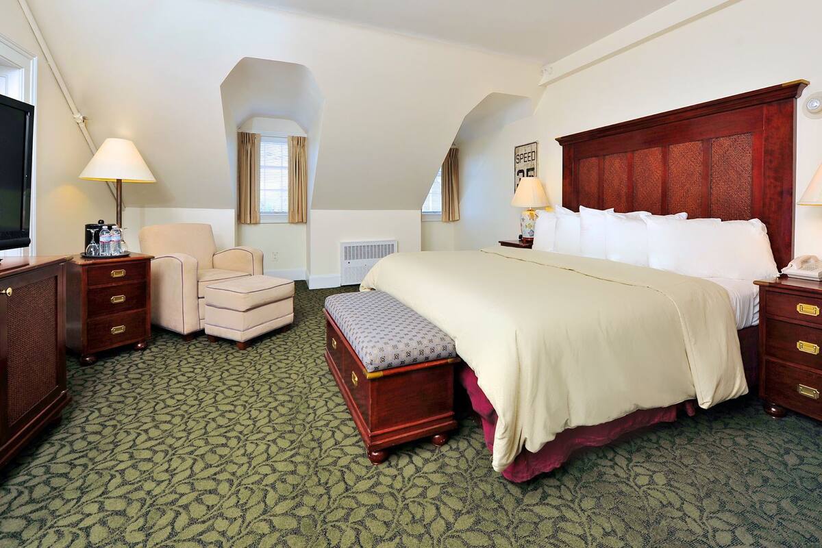 Une chambre d’hôtel avec un grand lit, un bureau avec une lampe, une chaise, une petite table avec une lampe, un téléviseur et une fenêtre avec des rideaux.