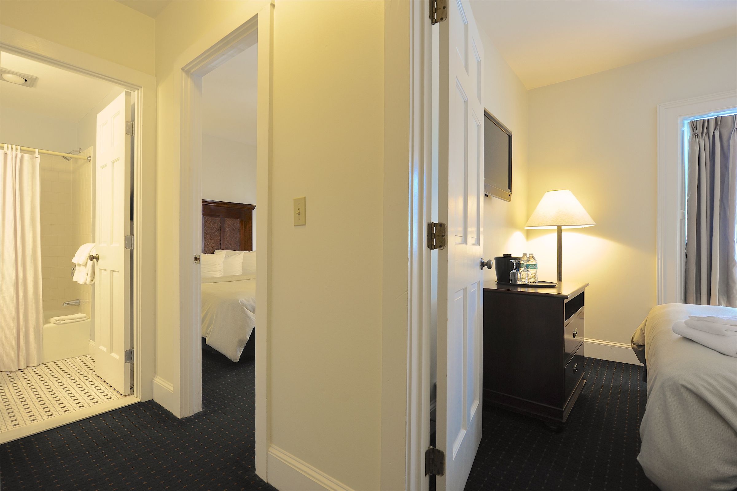 Habitación de hotel con cama, mesita de noche con lámpara, baño con ducha y puerta que da a otra habitación.