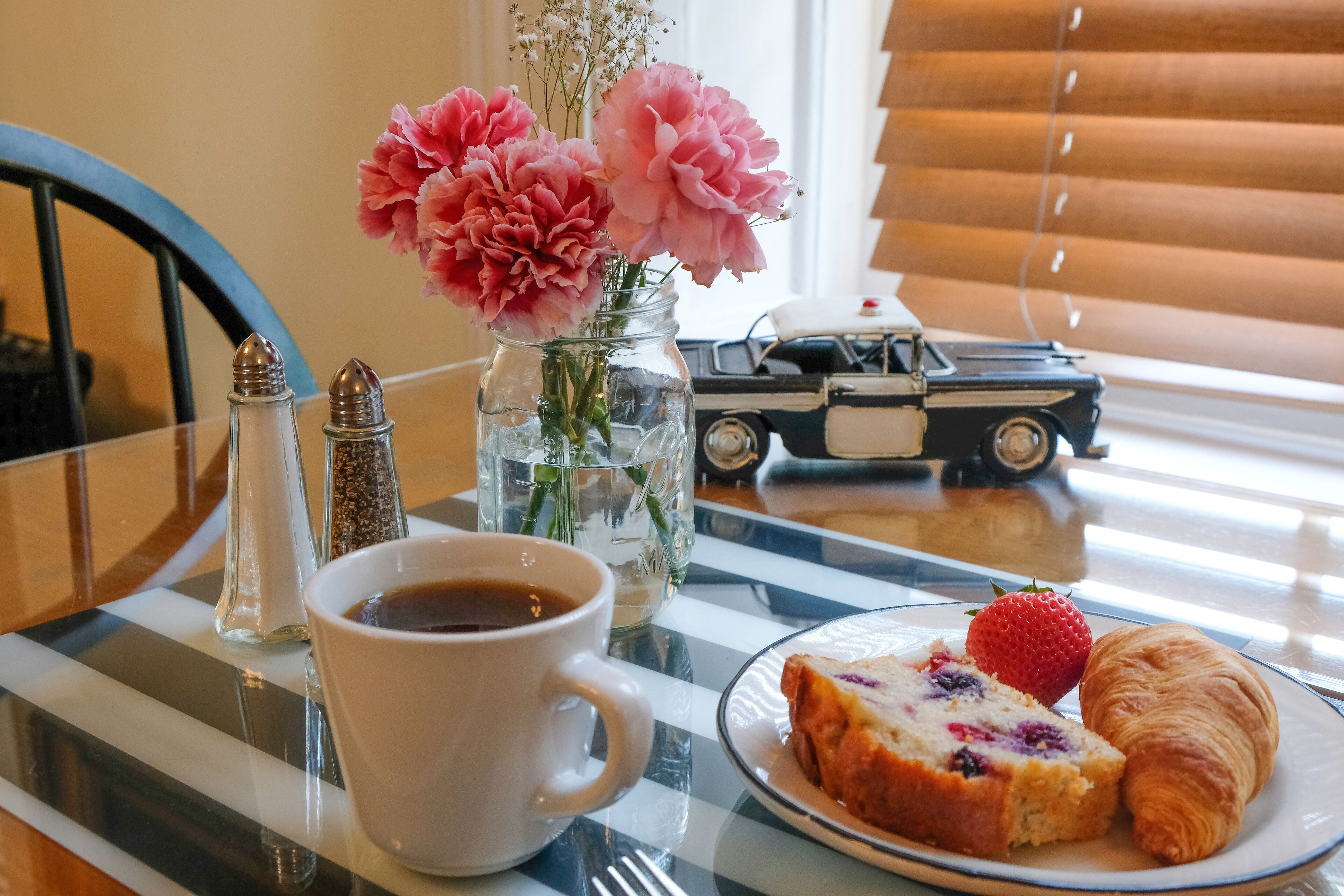 Uma mesa com uma xícara de café, um prato com um croissant e uma morango, um vaso de flores e um saleiro.