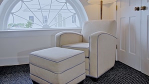 A room with a large arched window, a white armchair, a matching ottoman, a floor lamp, and a white door with a brass doorknob. - Jailhouse Inn (Newport)