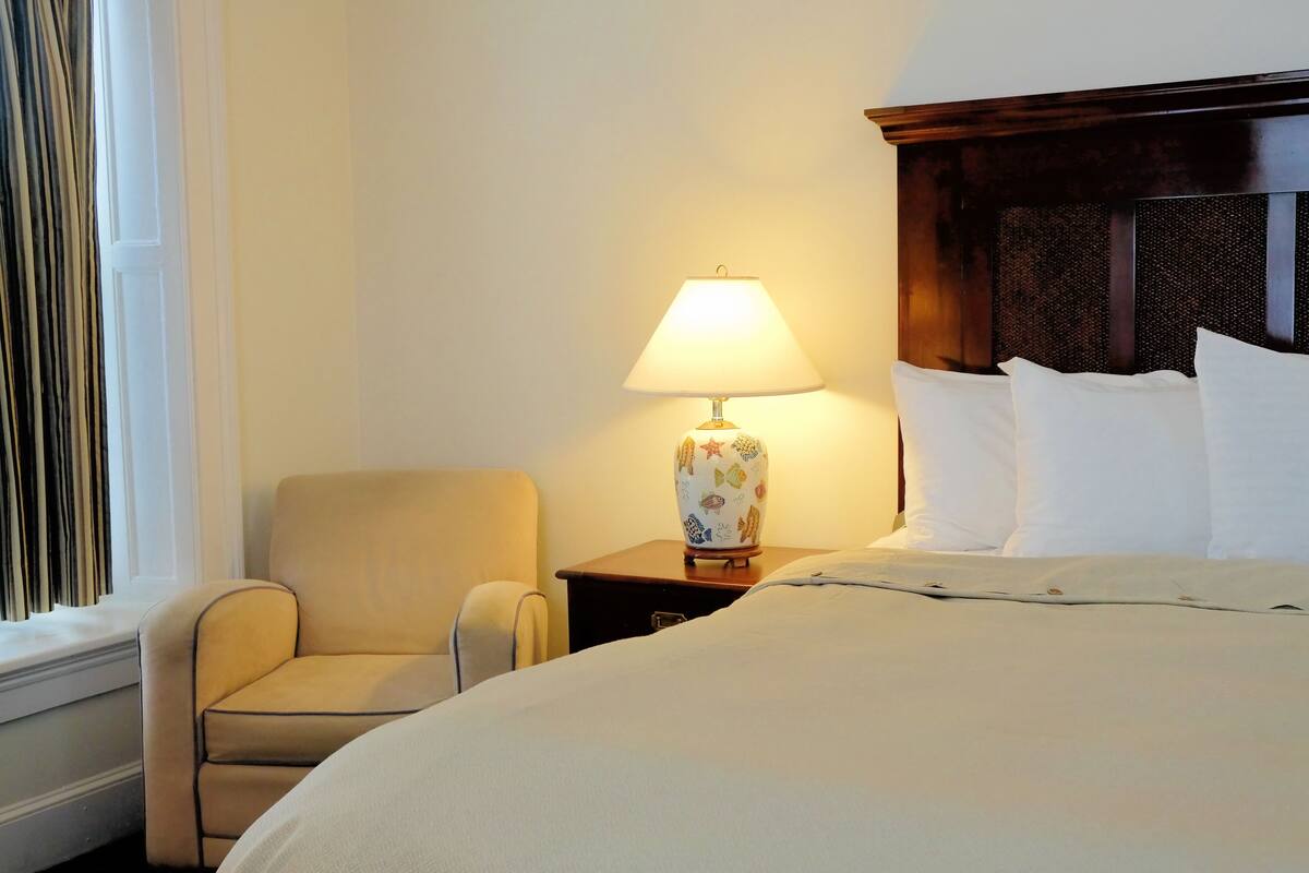 Une chambre d’hôtel avec un lit, une chaise, une table de chevet avec une lampe et une fenêtre avec des rideaux.