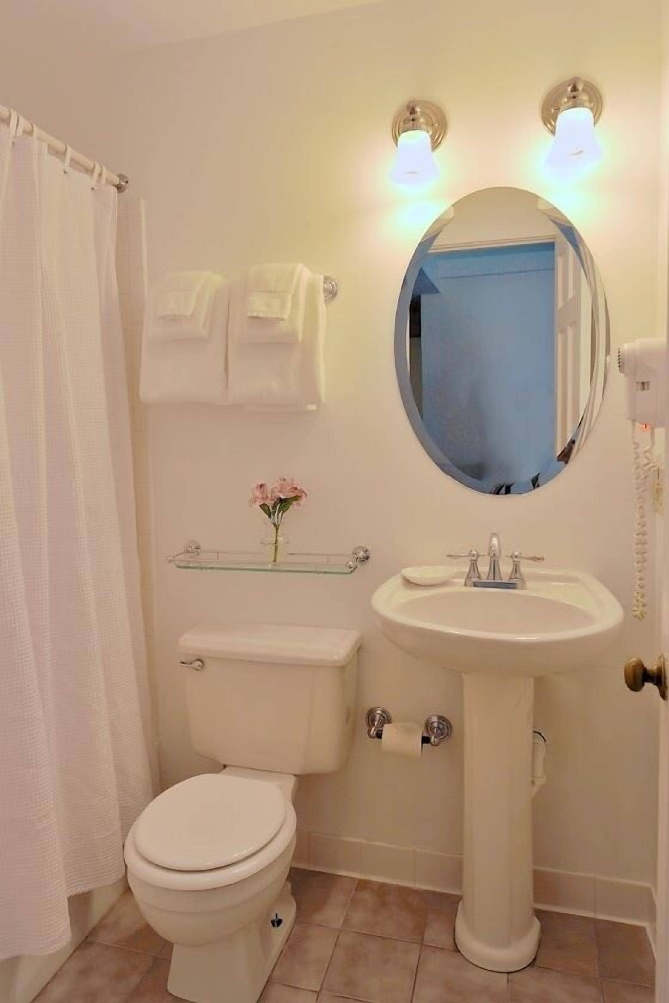 A bathroom with a toilet, sink, and mirror. There is a towel rack above the toilet and a glass shelf with a flower.