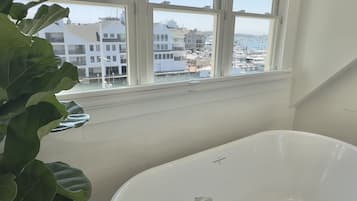 A bathroom with a bathtub, a window with a view of buildings, and a potted plant.