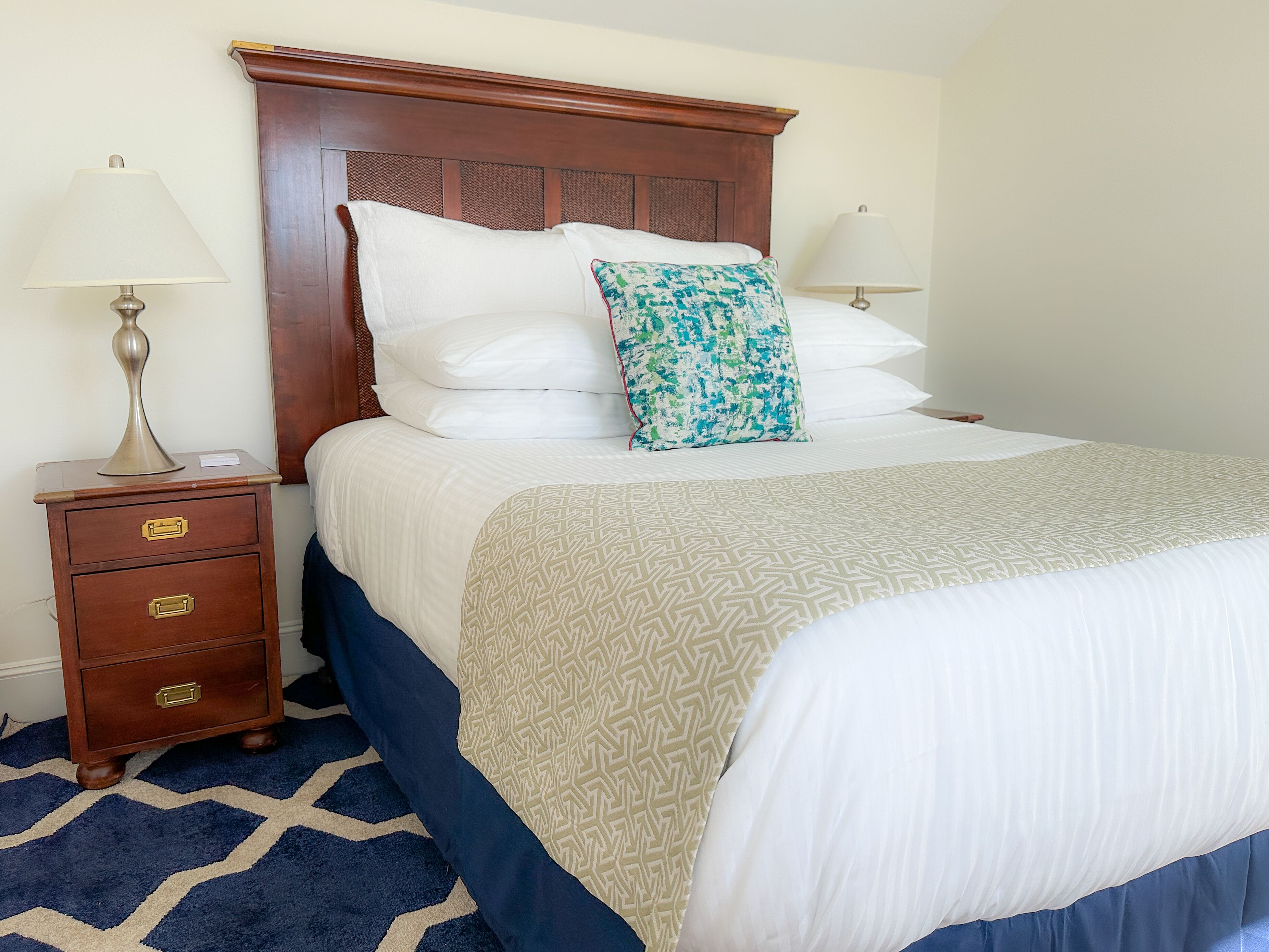 Cama bem arrumada com colcha estampada, mesa de cabeceira de madeira com gavetas e duas luminárias.