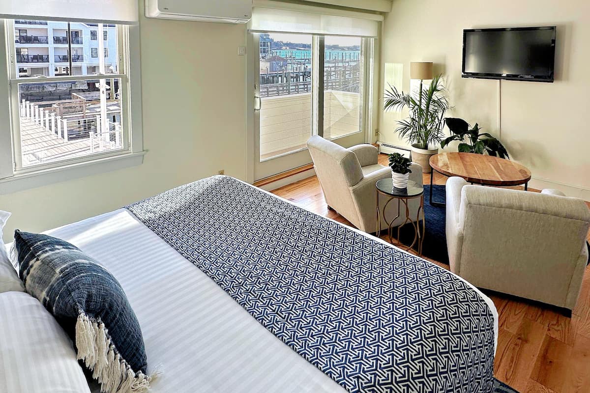 A hotel room with a bed, a chair, a small table, a TV, and a balcony with a view of buildings.