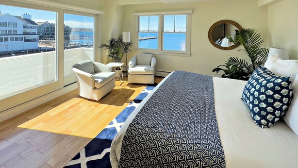 A spacious room with a large bed, two armchairs, a small table, and a view of the sea through large windows. - Harborside Inn (Newport)
