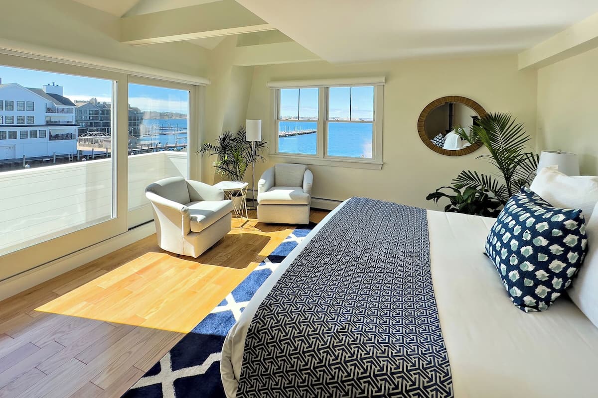 A spacious room with a large bed, two armchairs, a small table, and a view of the sea through large windows.