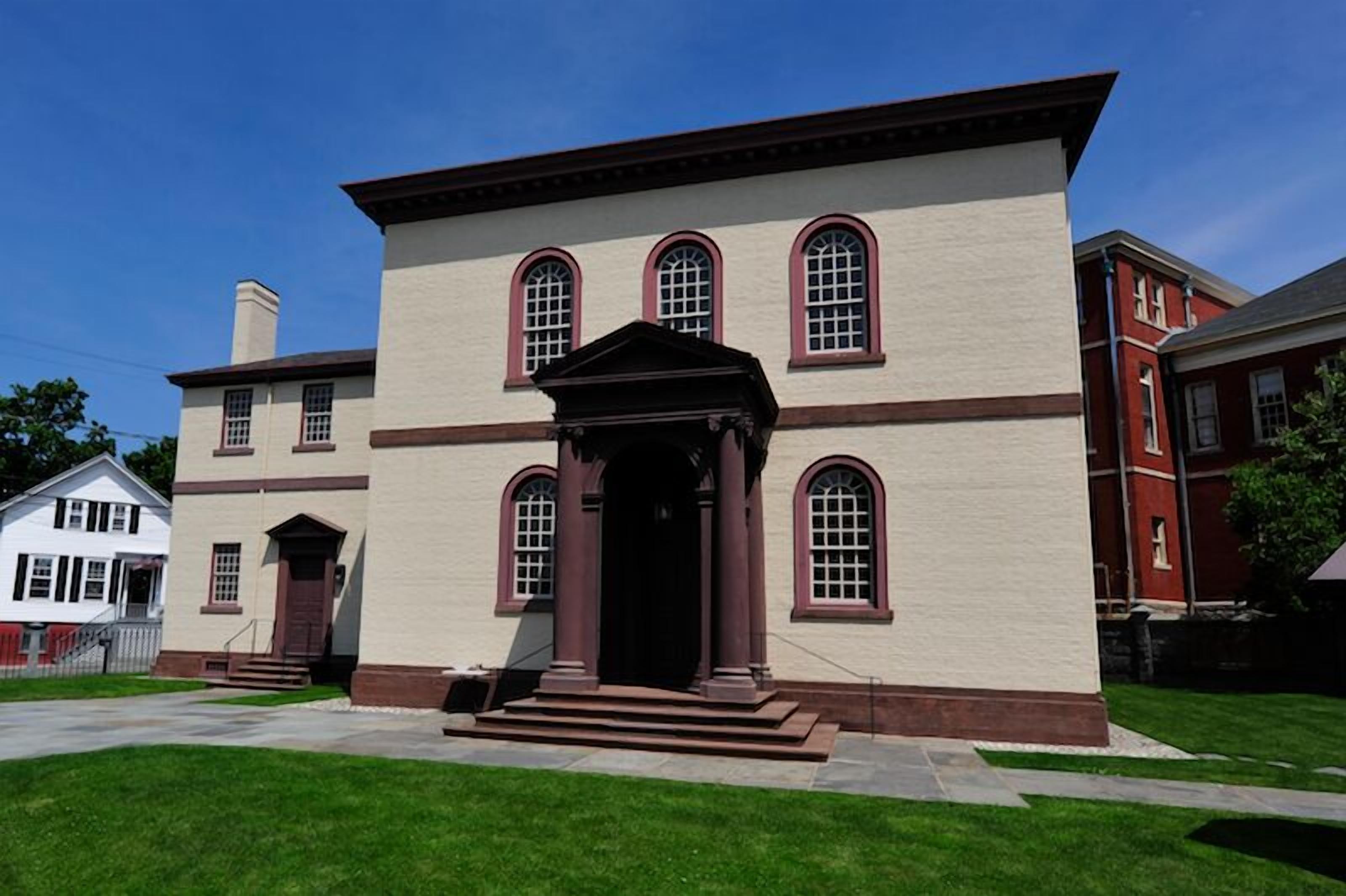 A two-story building with a central entrance flanked by columns, a symmetrical facade, and a red brick structure to the right.