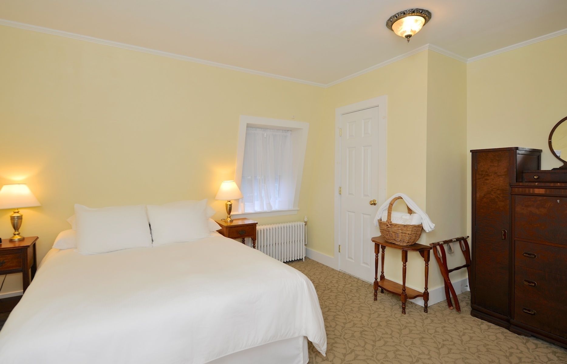 Une chambre d’hôtel avec un grand lit, deux lampes de chevet, un radiateur, une commode en bois et une petite table avec un panier.
