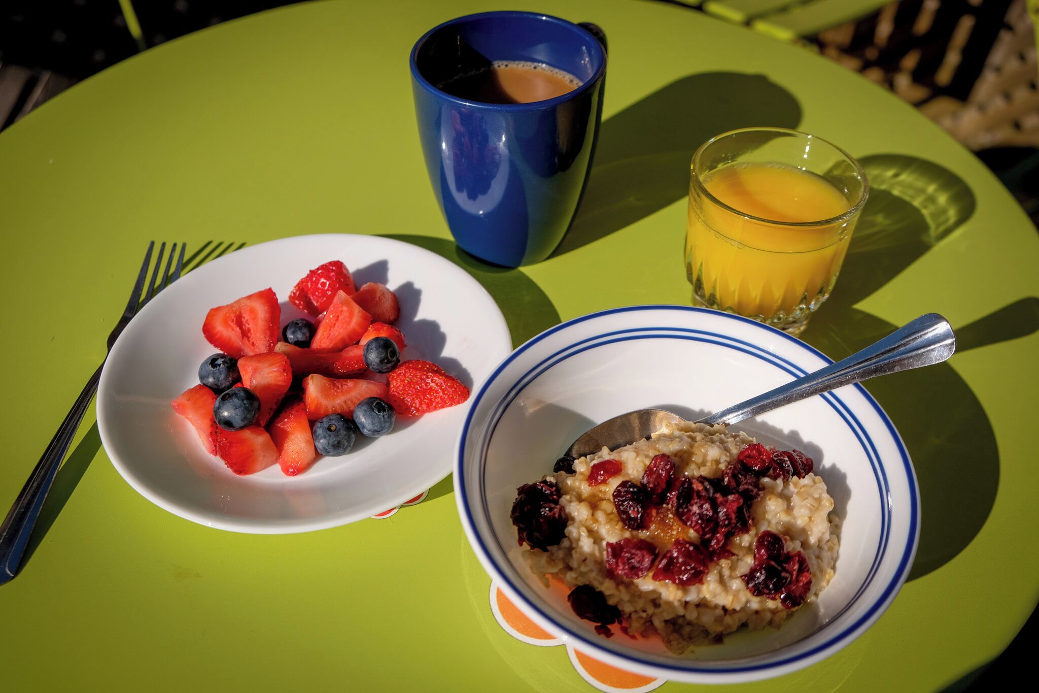 A breakfast setting with a bowl of oatmeal topped with berries, a plate of strawberries and blueberries, a glass of orange juice, and a blue mug.