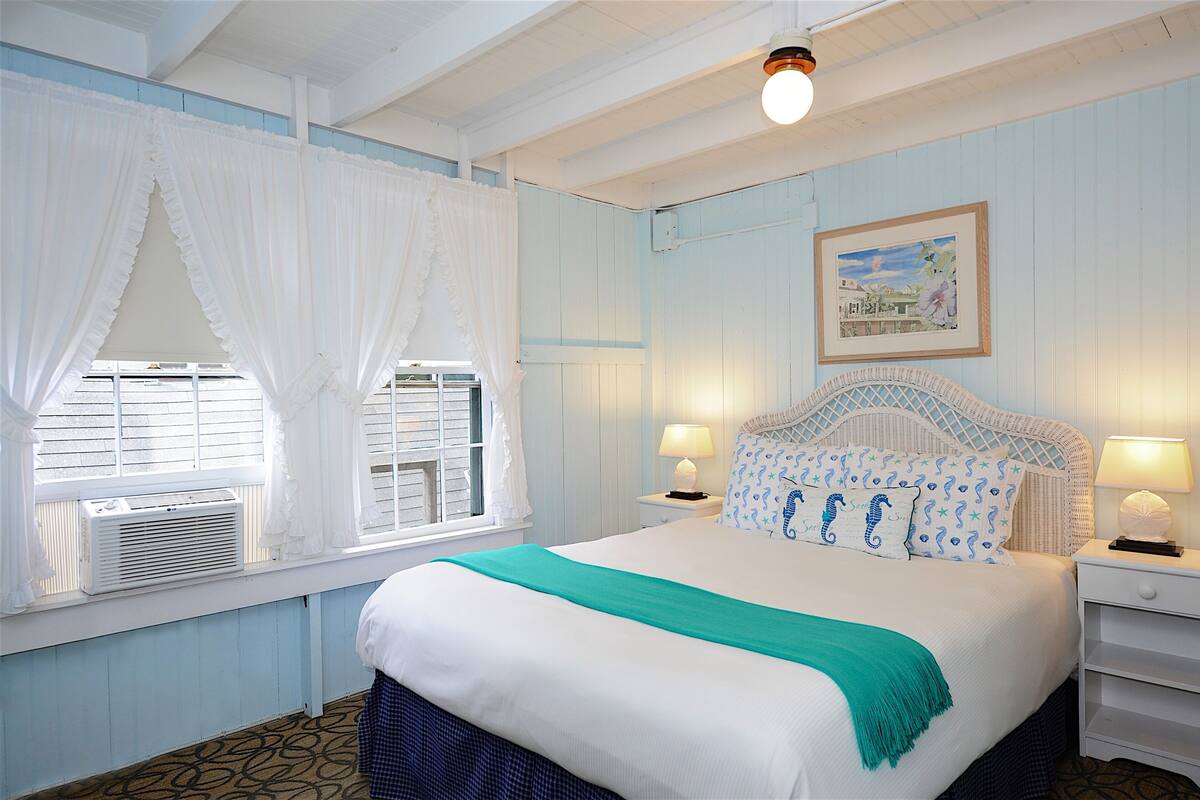 A bedroom with a bed, two bedside tables with lamps, a window with curtains, a wall-mounted air conditioner, and a framed picture on the wall.