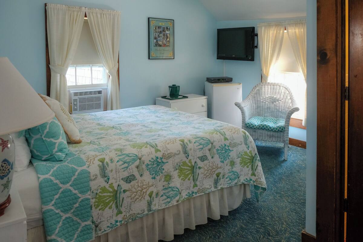 A bedroom with a bed, a chair, a television, a small table, and a window with curtains.