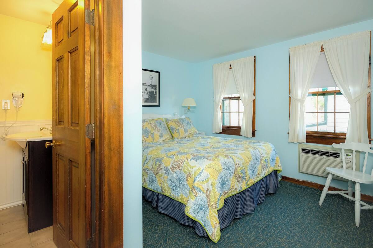 A bedroom with a bed, a chair, a window with curtains, a bedside lamp, and a wall-mounted air conditioning unit.