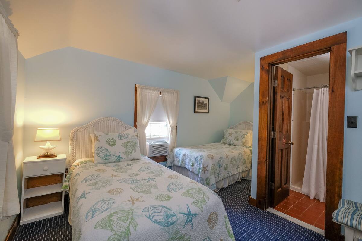 A bedroom with two beds, a nightstand, a window with curtains, a shower area, and a door leading to another room.