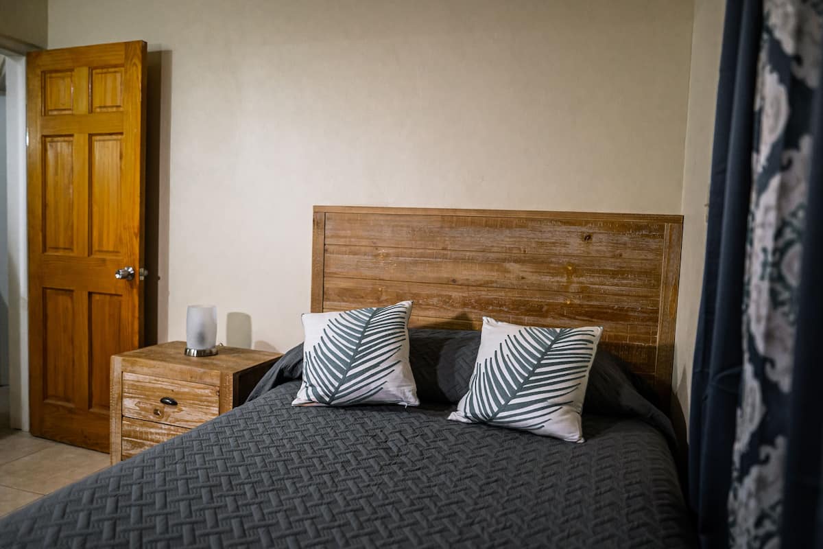 A bedroom with a wooden headboard, two pillows with leaf patterns, a bedside table with a lamp, and a wooden nightstand.