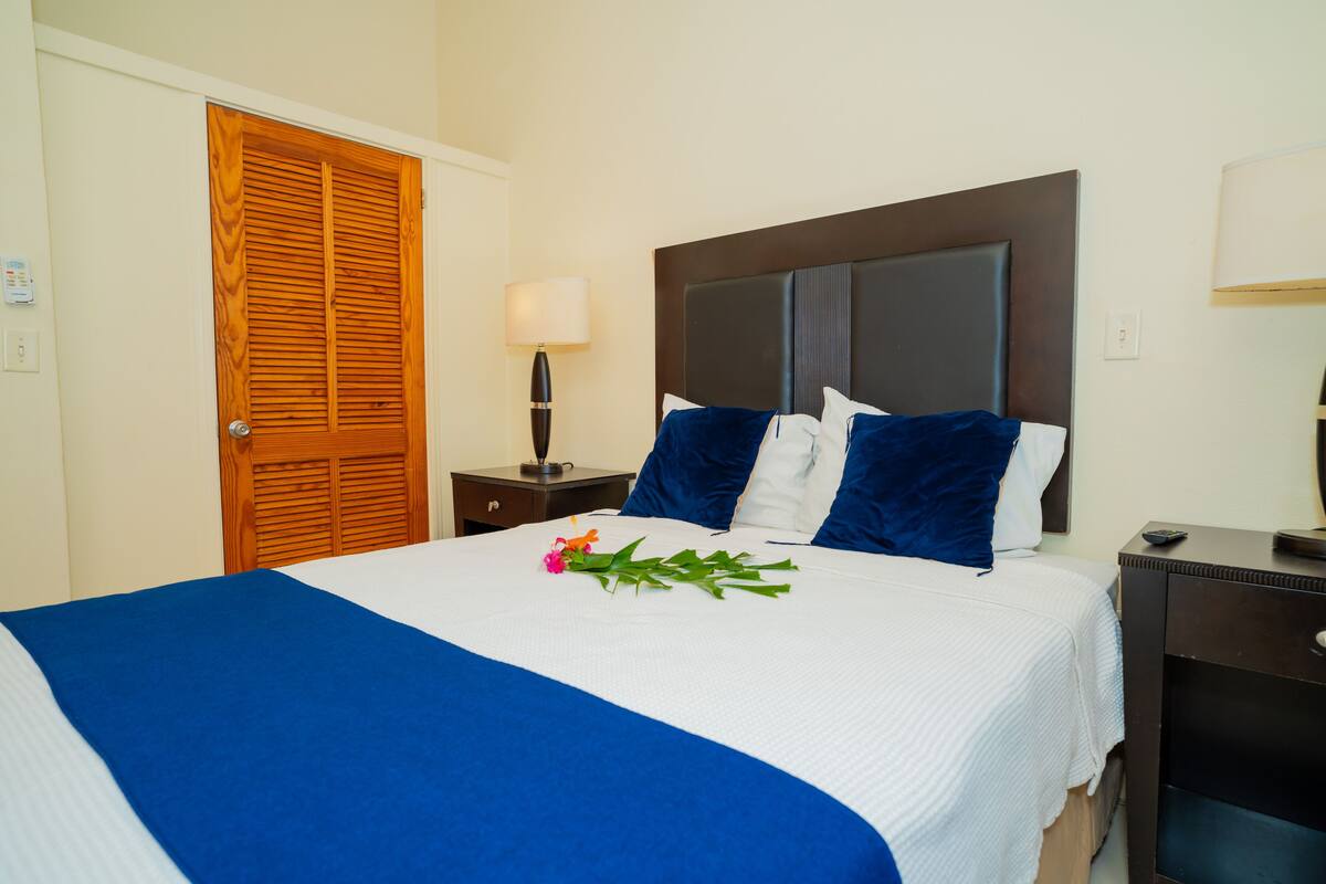 A hotel room with a large bed, a wooden door, a nightstand with a lamp, and a dresser.