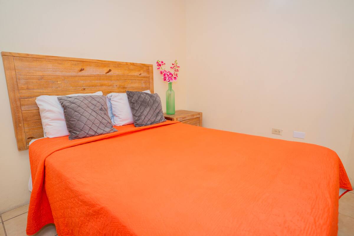 A bed with an orange bedspread, a wooden headboard, and a nightstand with a green vase and pink flowers.