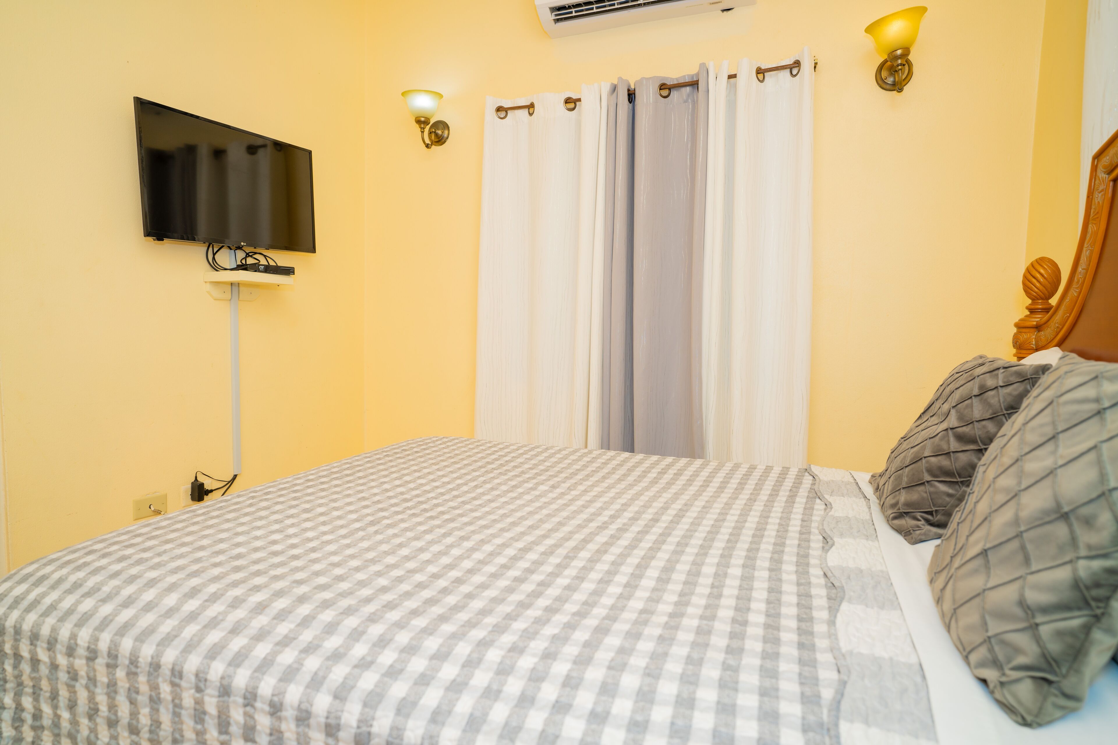 Un dormitorio con una colcha a cuadros, un televisor de pantalla plana en la pared, una cortina y dos lámparas fijadas a la pared.