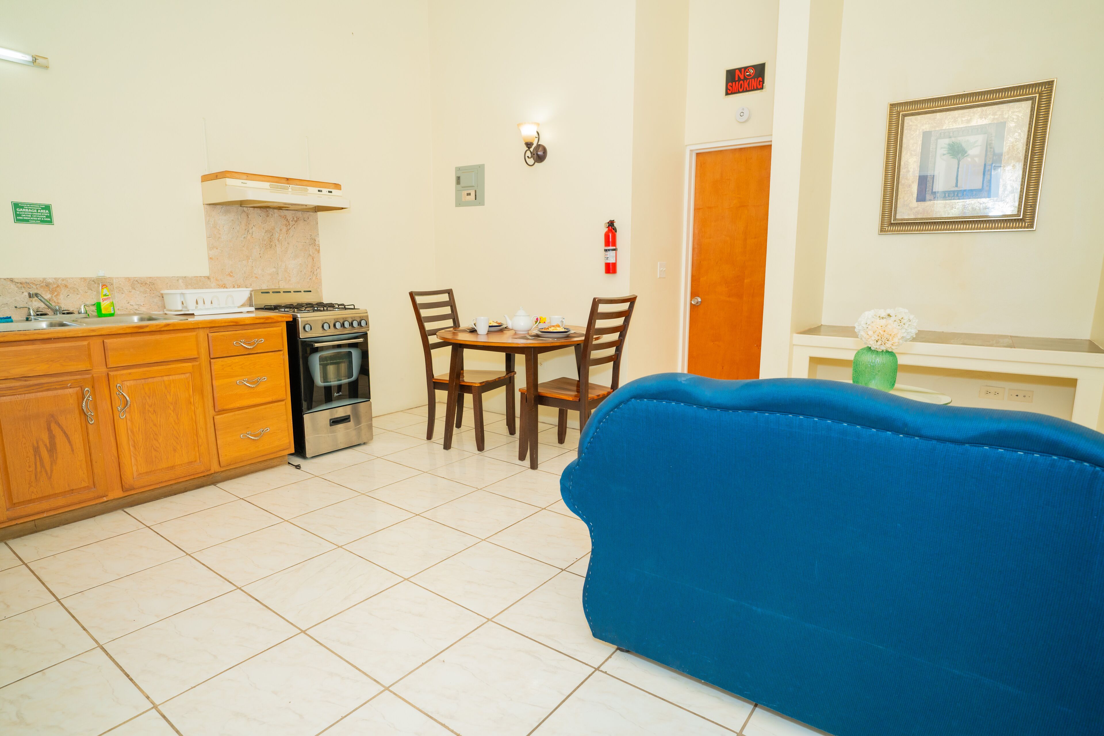 Una cocina compacta con estufa, pileta y muebles bajos de madera. Hay una mesa de comedor con sillas y un sofá azul en la sala.