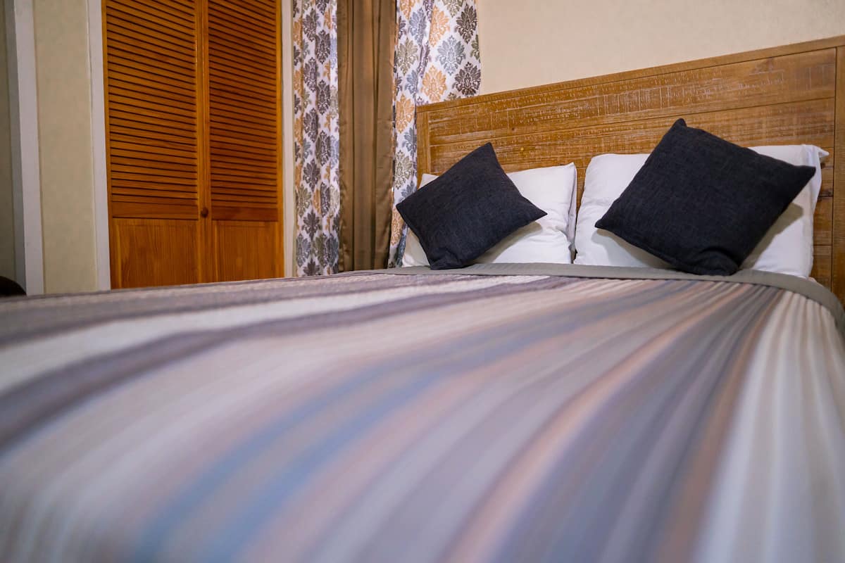 A bed with a striped bedspread and two pillows, a wooden headboard, and a patterned curtain.