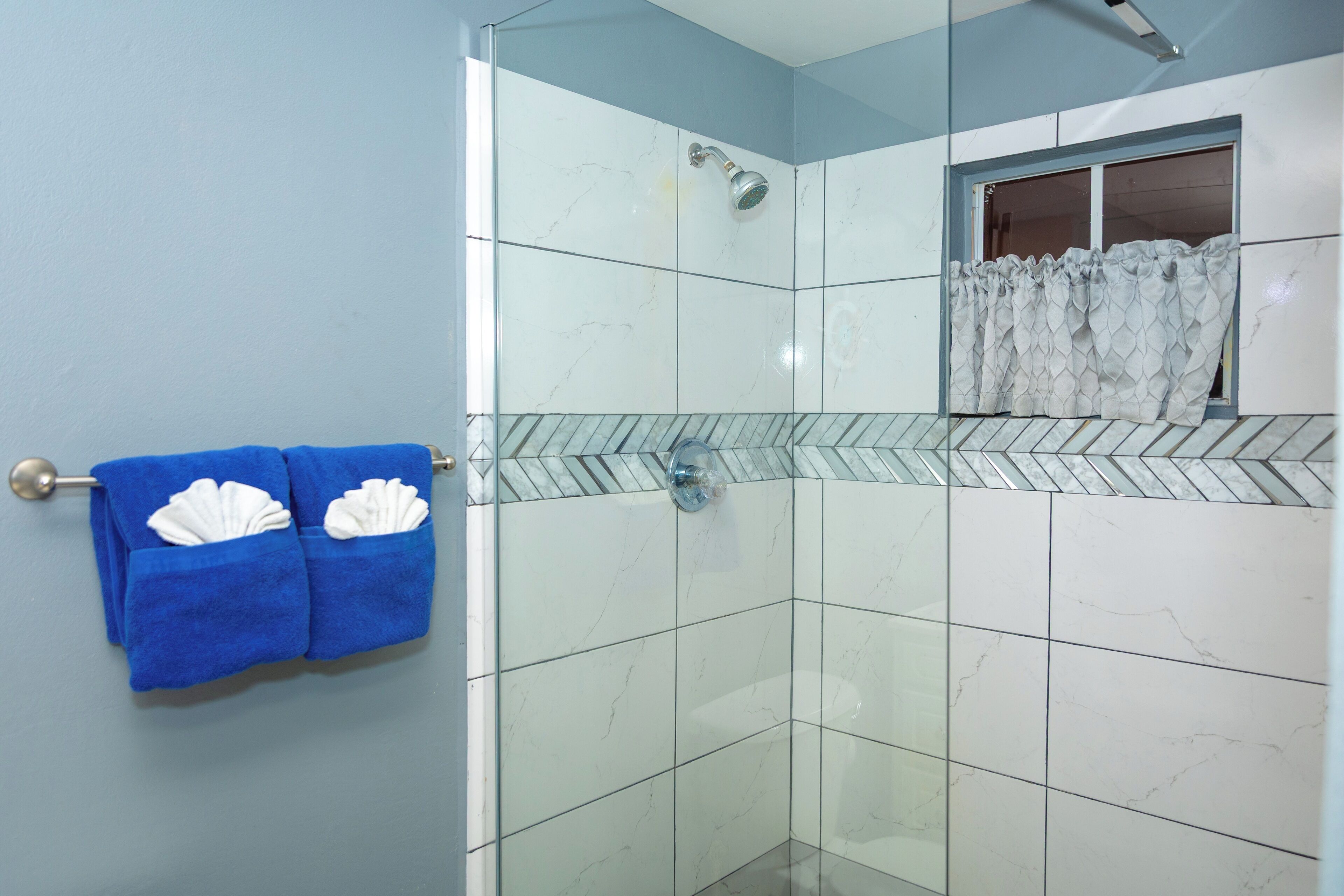 Une douche fermée par des parois de verre, dotée d’une fenêtre, recouverte de carreaux de marbre blanc et agrémentée d’une serviette bleue ornée de motifs à coquilles blanches.