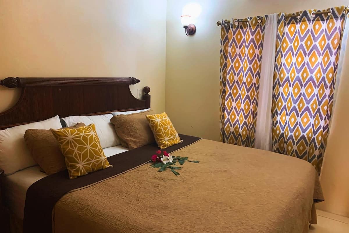 A neatly made bed with a patterned bedspread and pillows, a wooden headboard, and a wall-mounted light fixture.