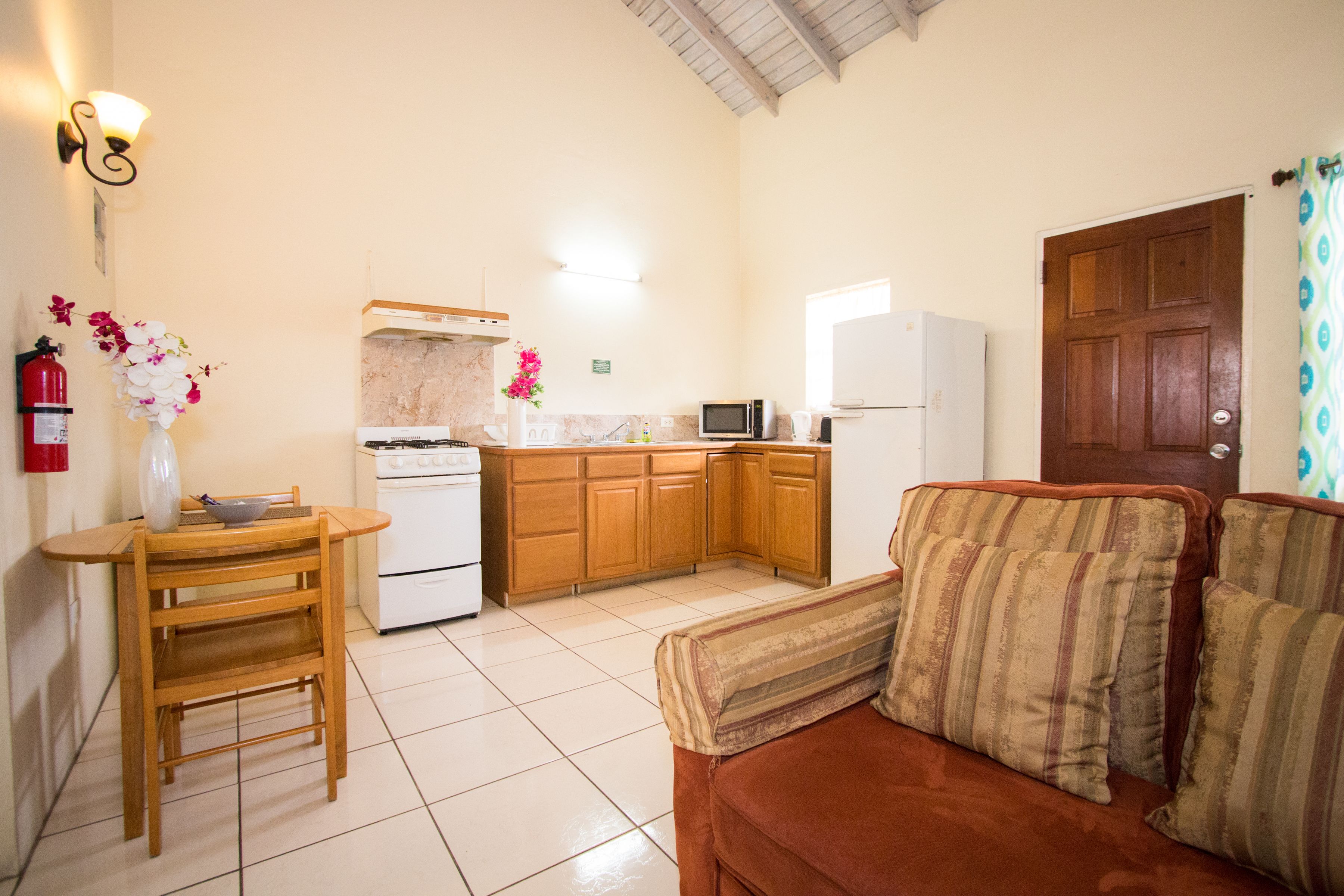 A compact kitchen with wooden cabinets, a microwave, and a stove. There is a dining table with a vase of flowers and a chair. A brown sofa with striped cushions is in the living area.