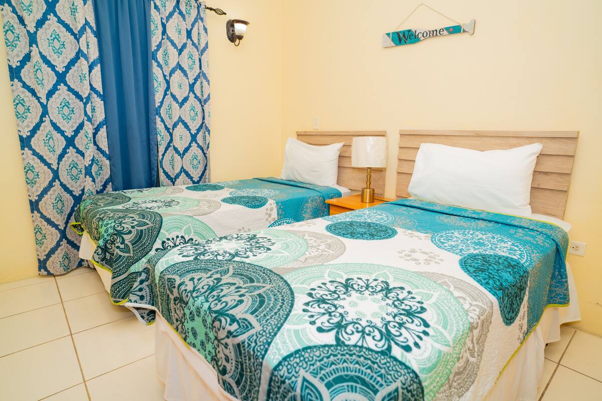 A room with two beds, a blue and white patterned curtain, a 'Welcome' sign, and a wall-mounted lamp.