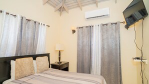 A hotel room with a bed, curtains, a nightstand with a lamp, an air conditioning unit, and a mounted television. - Connie's Comfort Suites (St. John's)