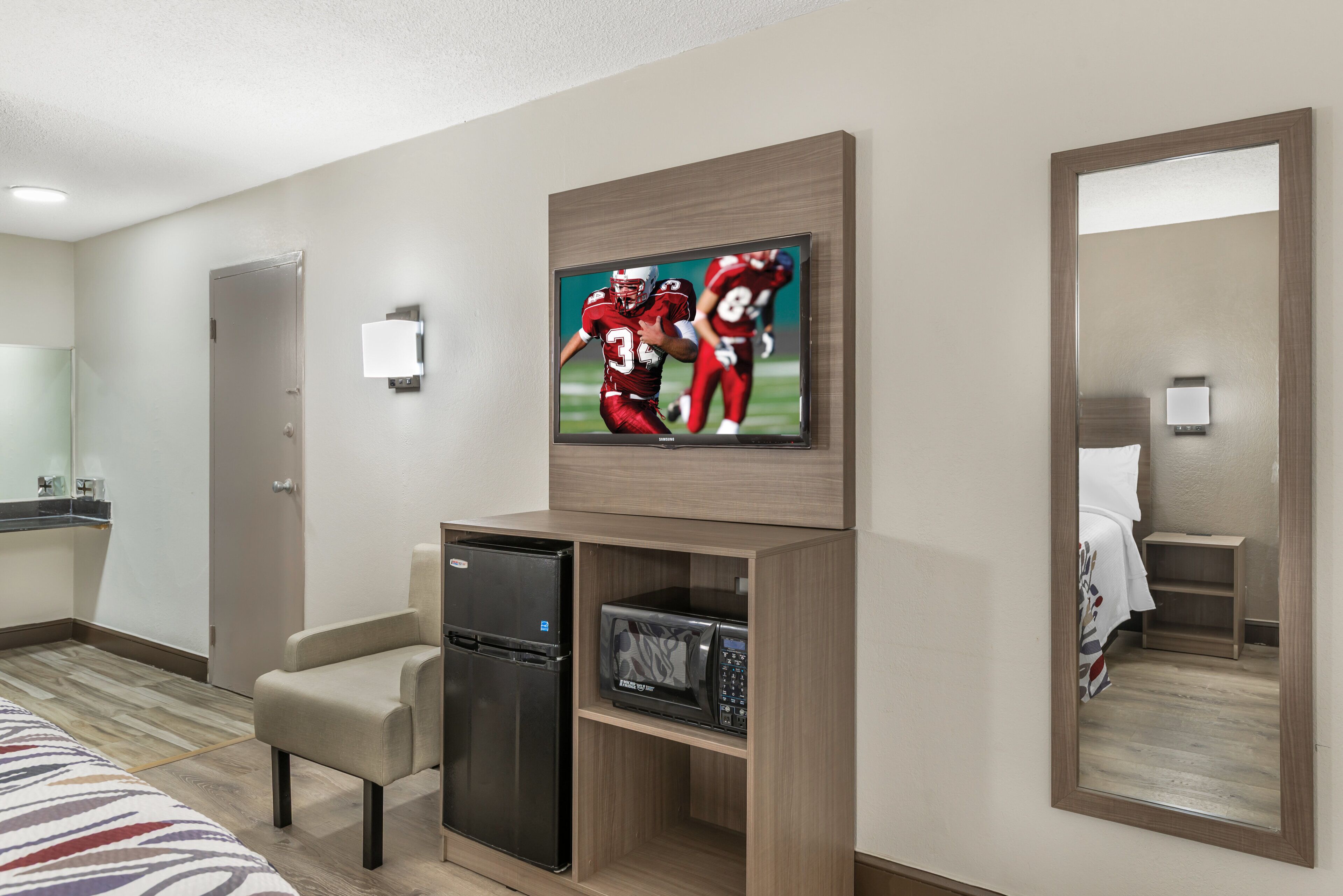 Habitación de hotel con televisión de pantalla plana en la pared, un mini refrigerador, microondas, una zona de estar con una silla y un espejo grande.