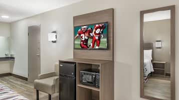 Habitación de hotel con televisión de pantalla plana en la pared, un mini refrigerador, microondas, una zona de estar con una silla y un espejo grande.