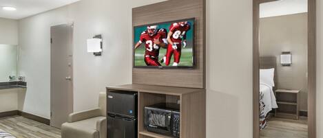 A hotel room with a flat-screen TV mounted on the wall, a mini-fridge, a microwave, a seating area with a chair, and a large mirror.