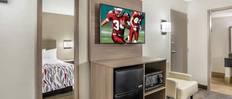 A hotel room with a bed, a TV mounted on the wall, a refrigerator, a microwave, and a seating area.