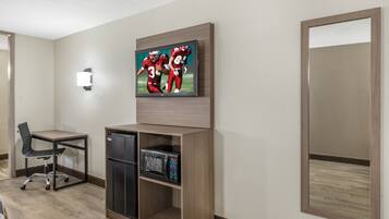 A hotel room with a flat-screen TV mounted on the wall, a compact refrigerator, a microwave, a wooden cabinet, a chair, and a large mirror.