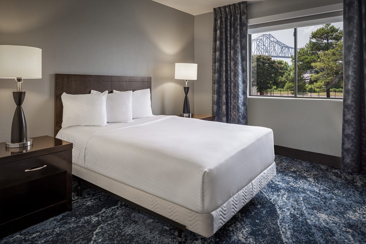 Una habitación de hotel con una cama grande, dos mesitas de noche, una ventana con vistas a un puente y una alfombra estampada.