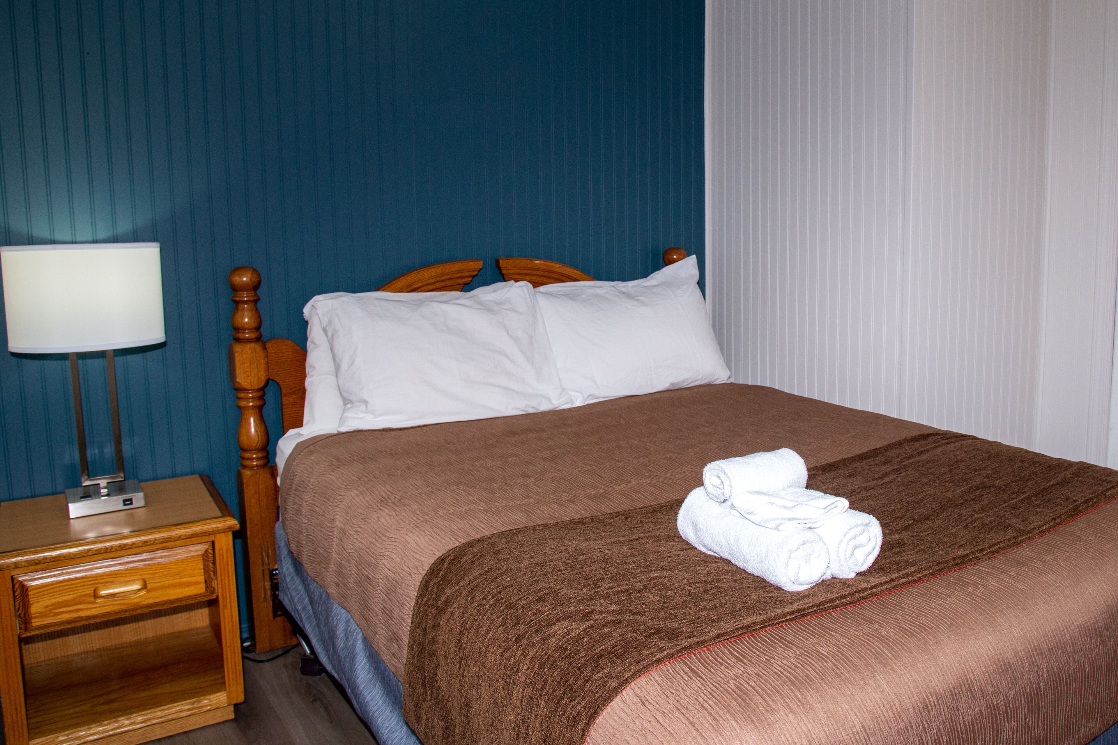 Un lit bien fait, avec une couverture marron et des oreillers blancs, une table de chevet en bois avec une lampe, et un mur bleu.
