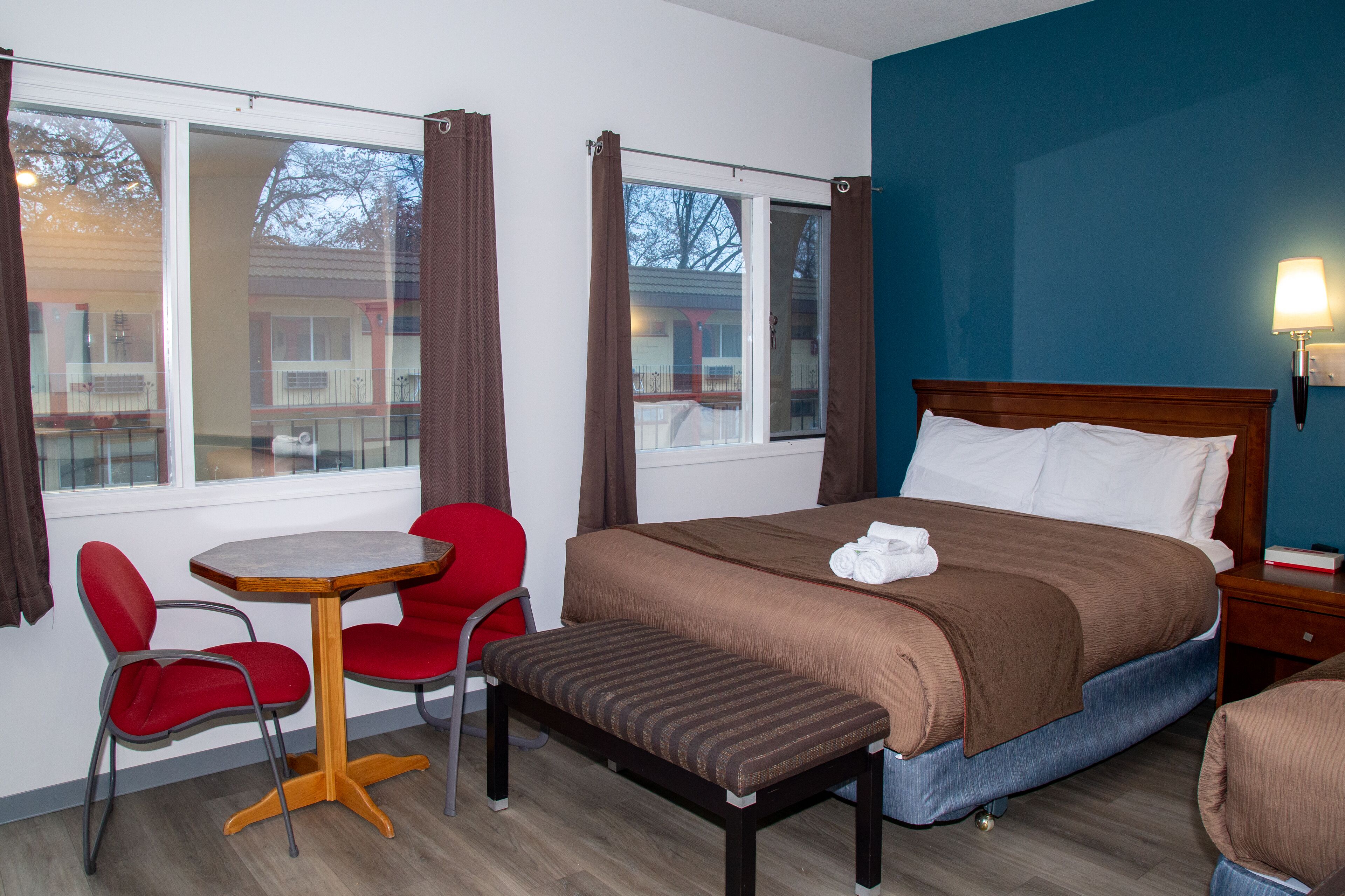 A hotel room with a bed, a small table, two red chairs, a bench, and a window with curtains.