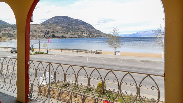 A view from a balcony with a metal railing, overlooking a body of water, a beach, and a hilly landscape. - Spanish Villa Resort (Penticton)