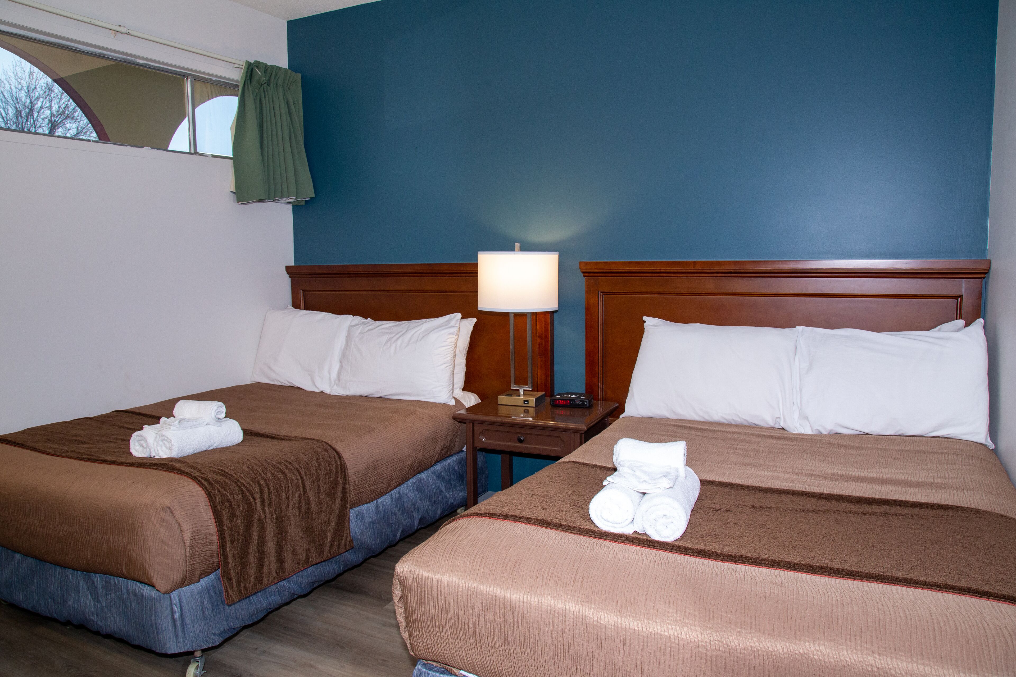 Two double beds with brown bedding, a nightstand with a lamp, and a window with a curtain.